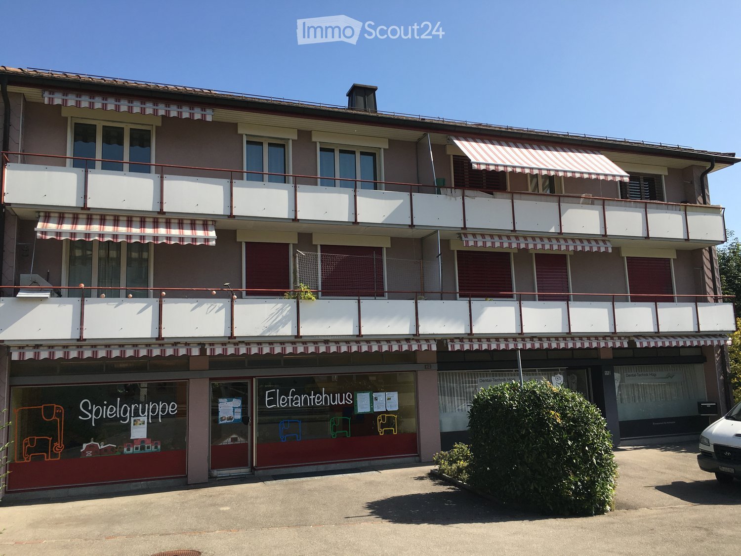 Three-story building with multiple balconies, striped awnings, and storefronts including 'Spielgruppe' and 'Elefantenhaus'.