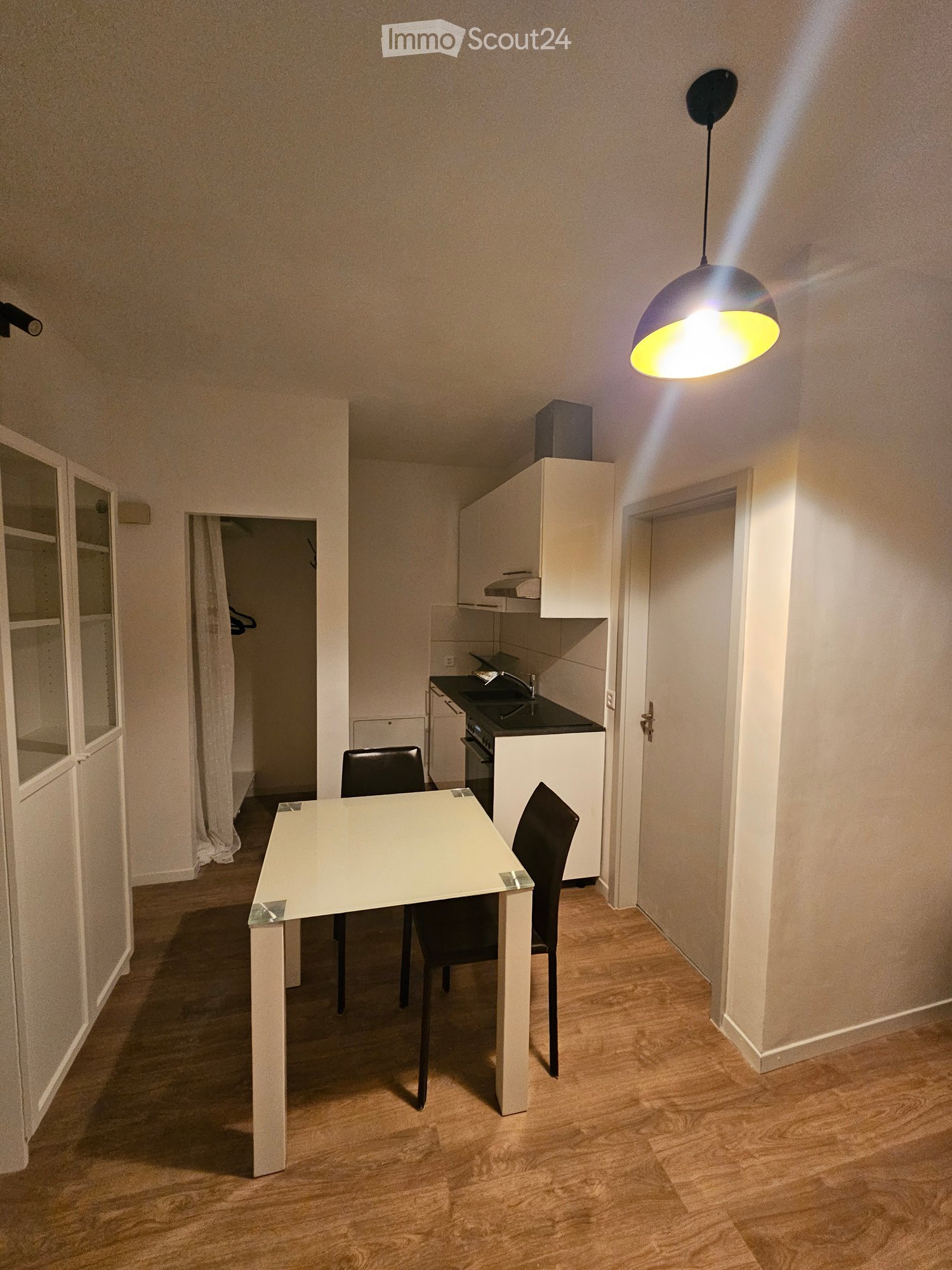 A modern, well-lit room with a white dining table and two black chairs, a small kitchen area with cabinets, an oven, and a sink, and a hanging light fixture.