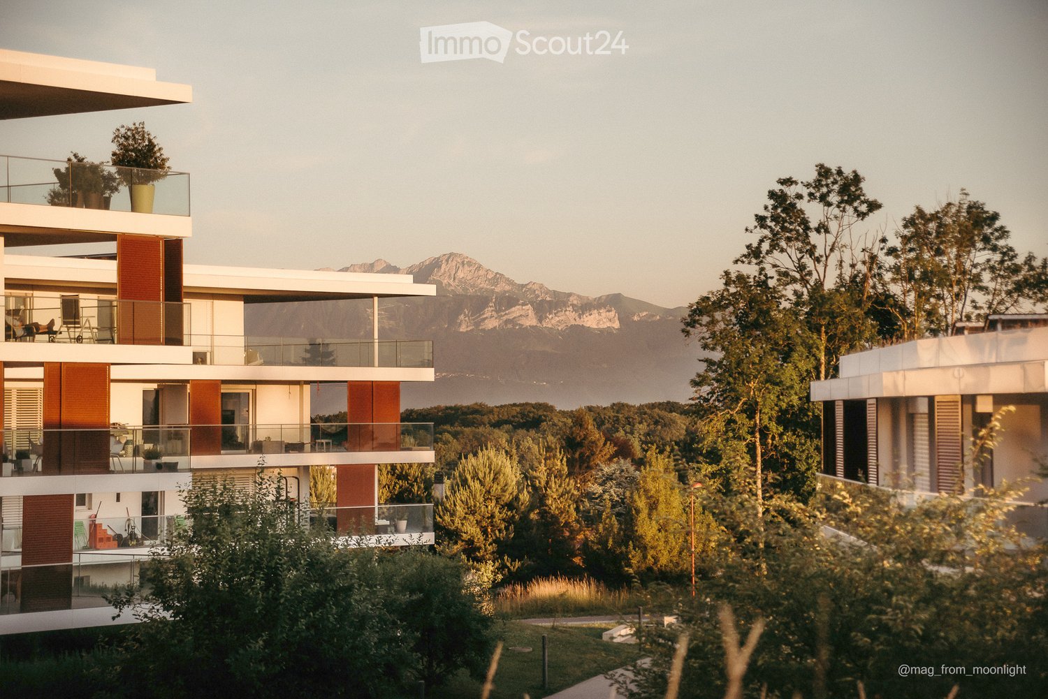 Modern multi-story residential building with balconies, large windows, and a scenic mountain view in the background.