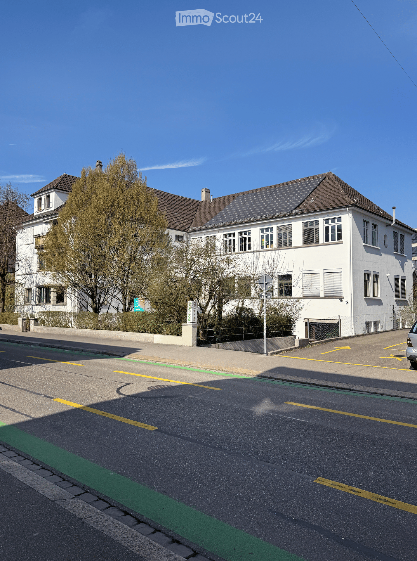 Two-story white building with multiple windows and a brown roof, featuring solar panels on the roof and surrounded by trees and a paved road.