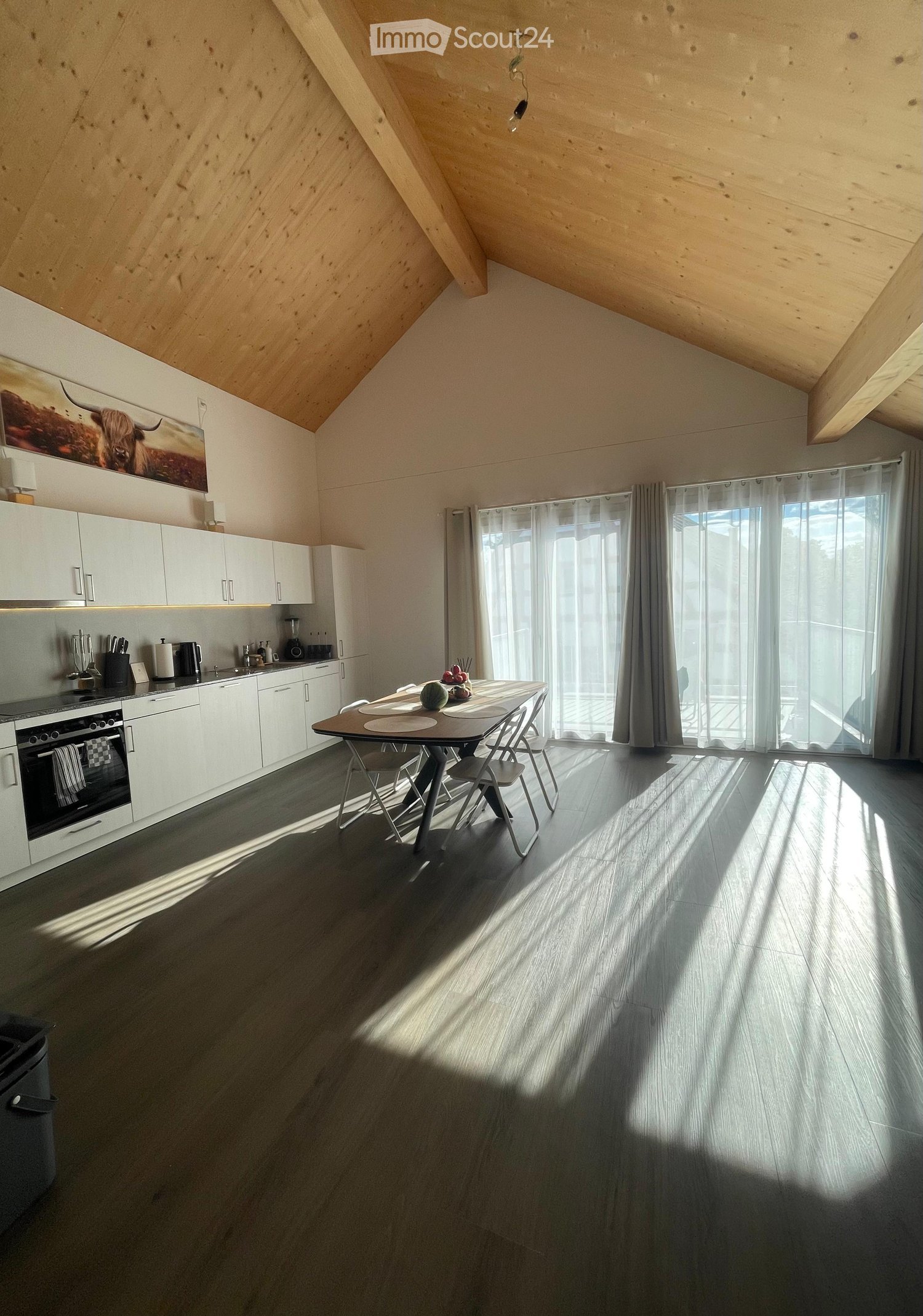 Modern kitchen with white cabinetry, stainless steel appliances, a dining table with chairs, and large windows with sheer curtains.