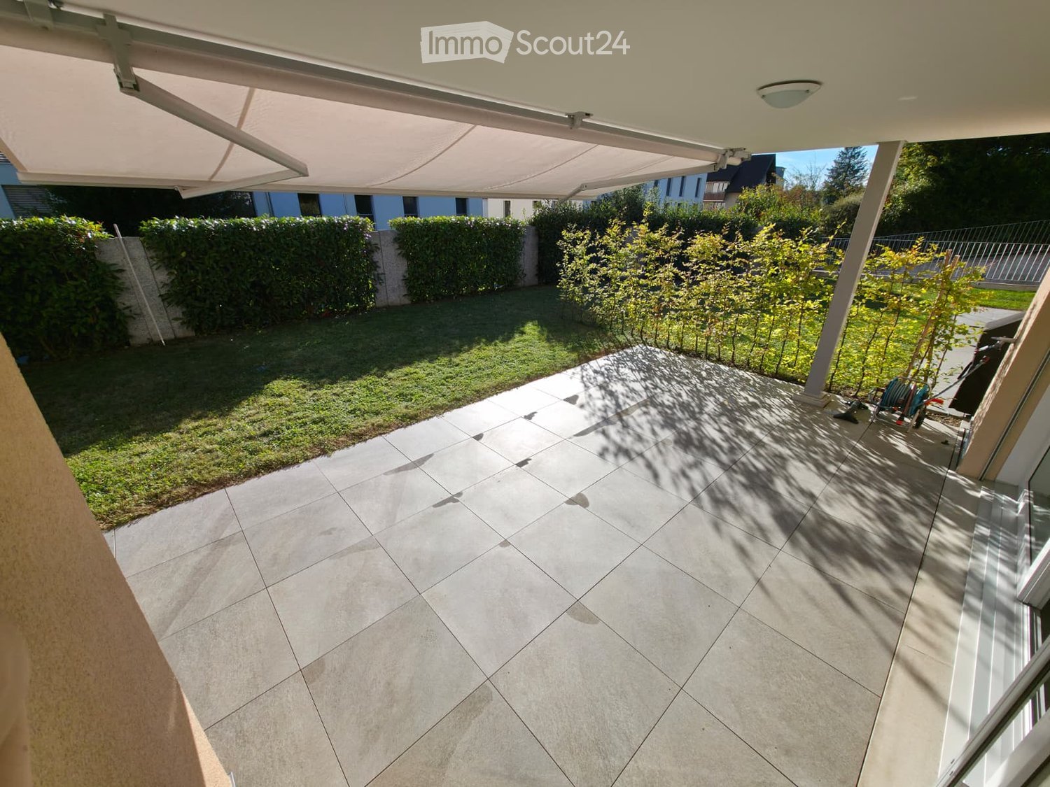 A well-maintained garden area with a tiled patio, covered by a retractable awning, surrounded by neatly trimmed hedges and small trees.