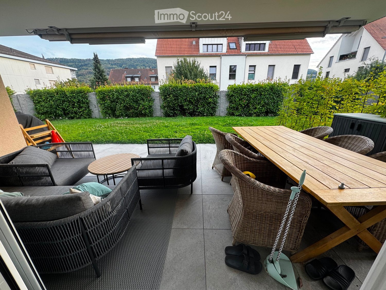 Balcony with a sofa, small table, chairs, and a larger wooden dining table with chairs, surrounded by greenery and a view of neighboring houses.