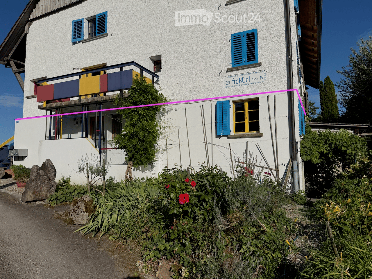 Two-story white building with blue shutters, a balcony, and a garden with flowers and plants.