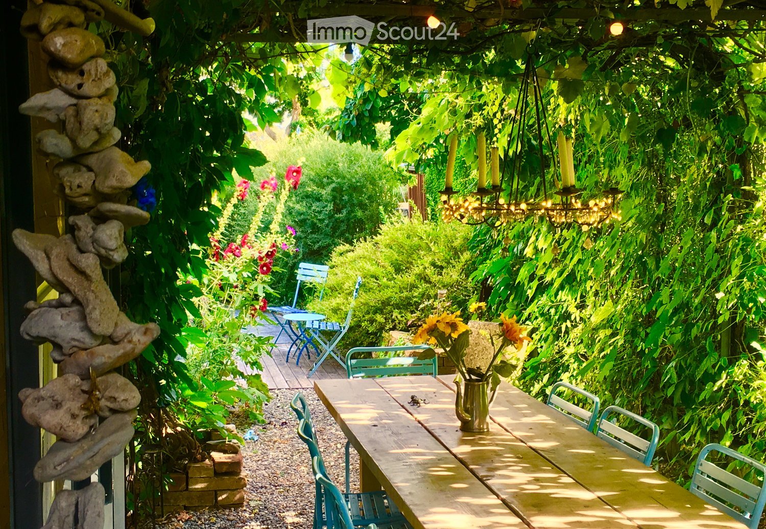 A lush garden area with a wooden table, chairs, hanging chandelier, and colorful flowers, surrounded by greenery and a stone sculpture.