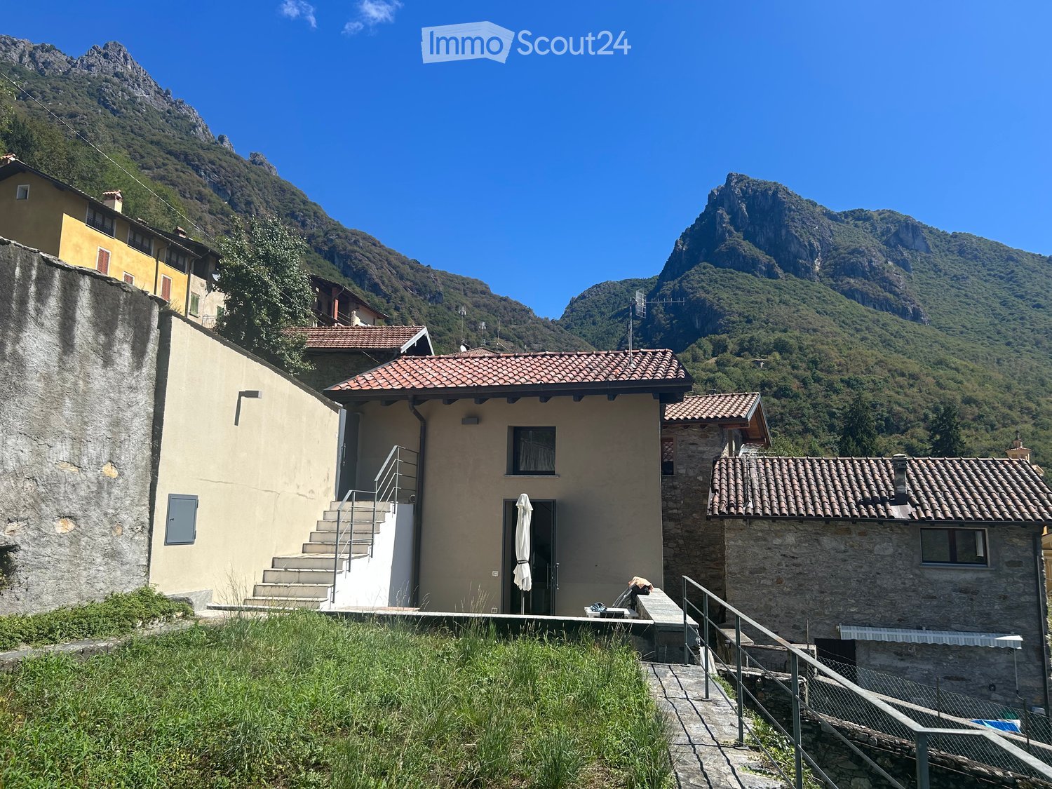 A two-story house with a tiled roof, stone and plaster exterior, and a small porch with an umbrella. The house is situated in a mountainous area with a staircase leading up to the entrance.