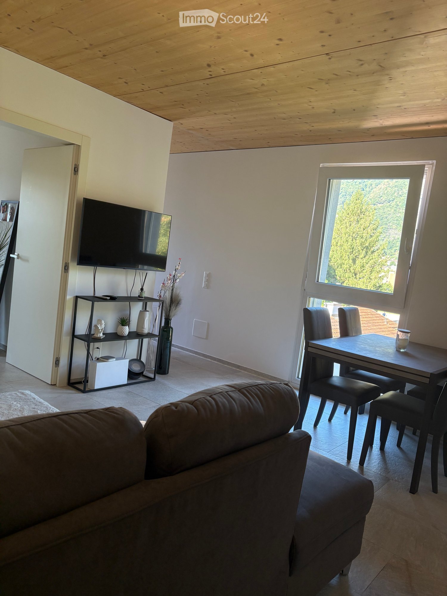 Modern living room with a brown leather sofa, a black dining table with four chairs, a television on a stand, a shelf with decorative items, and a large window with a view of greenery.
