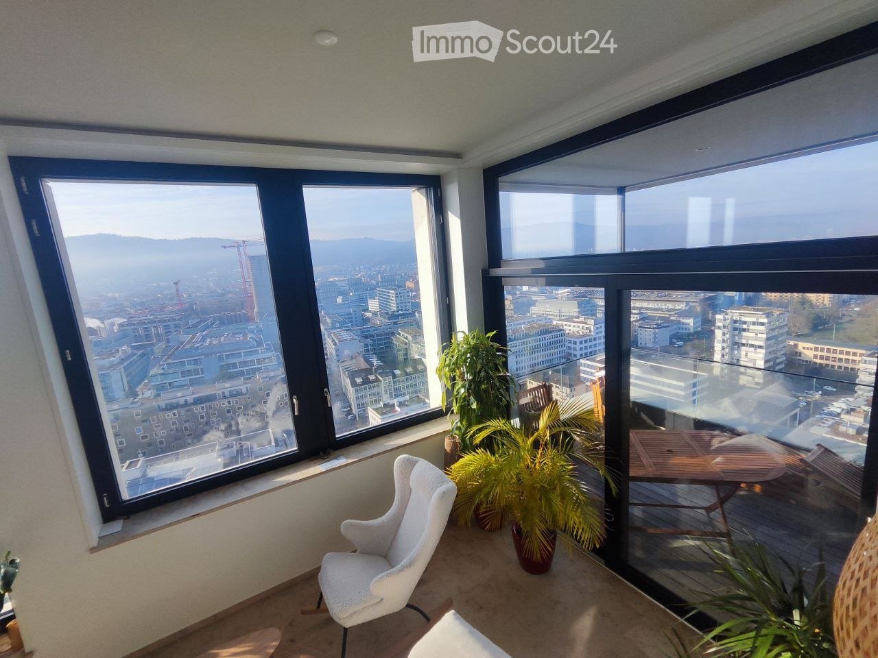 Bright room with large windows offering a cityscape view, furnished with a white chair, a small table, and several potted plants.