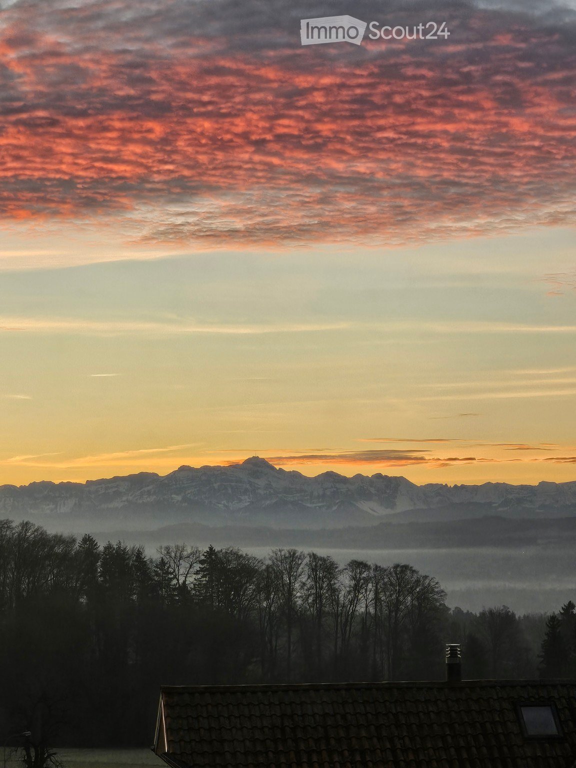 A serene view from a rooftop terrace showcasing a picturesque mountain range at sunset, with a clear sky transitioning from warm orange to deep blue hues, surrounded by silhouettes of trees and a distant forest.
