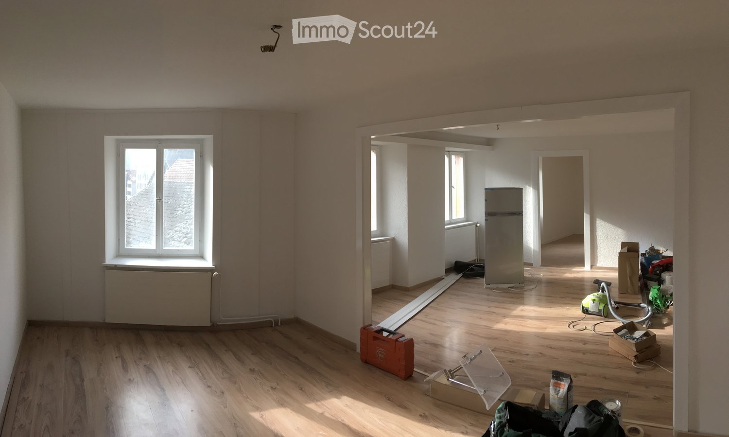 Empty room with wooden flooring, white walls, and large windows. Some construction materials and tools are visible, indicating ongoing renovation.