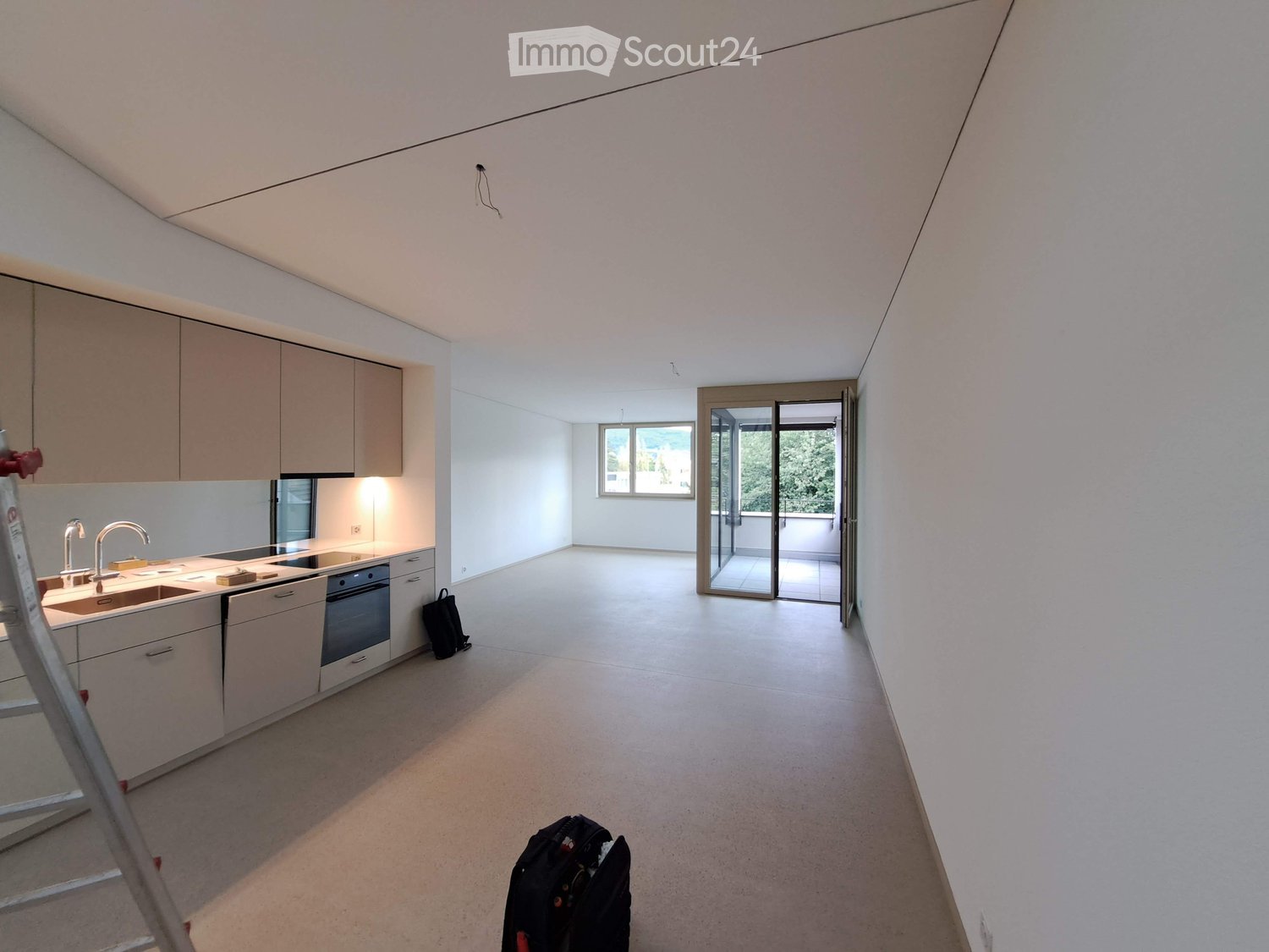 Modern kitchen with white cabinets, stainless steel sink, and built-in oven. Open space with large windows and sliding glass doors leading to a balcony.