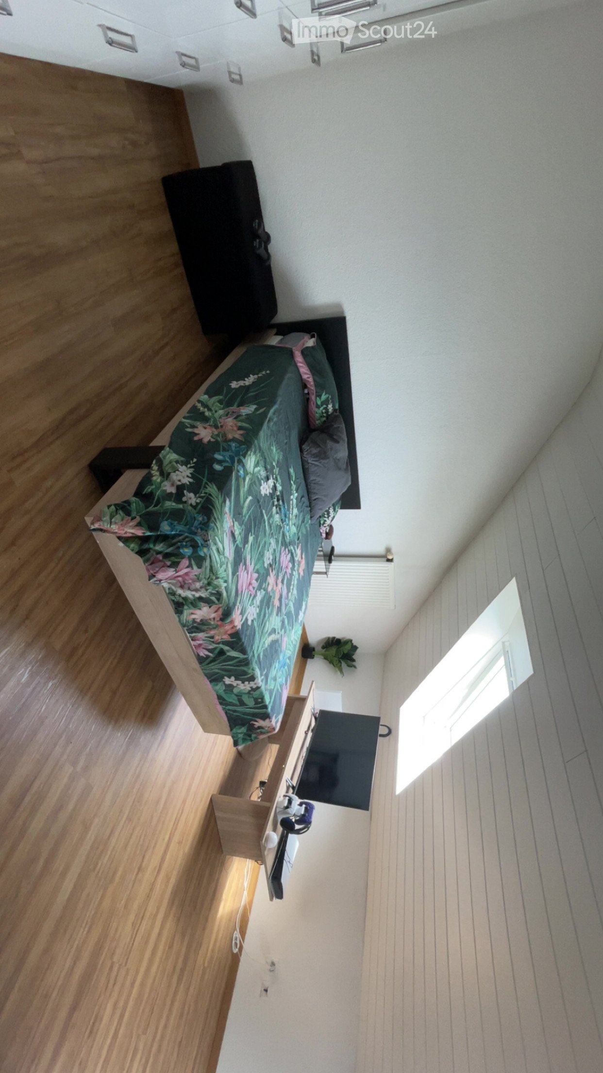 Modern bedroom with wooden flooring, a bed with a green and pink floral duvet, a nightstand with a television, and a small plant near the window.