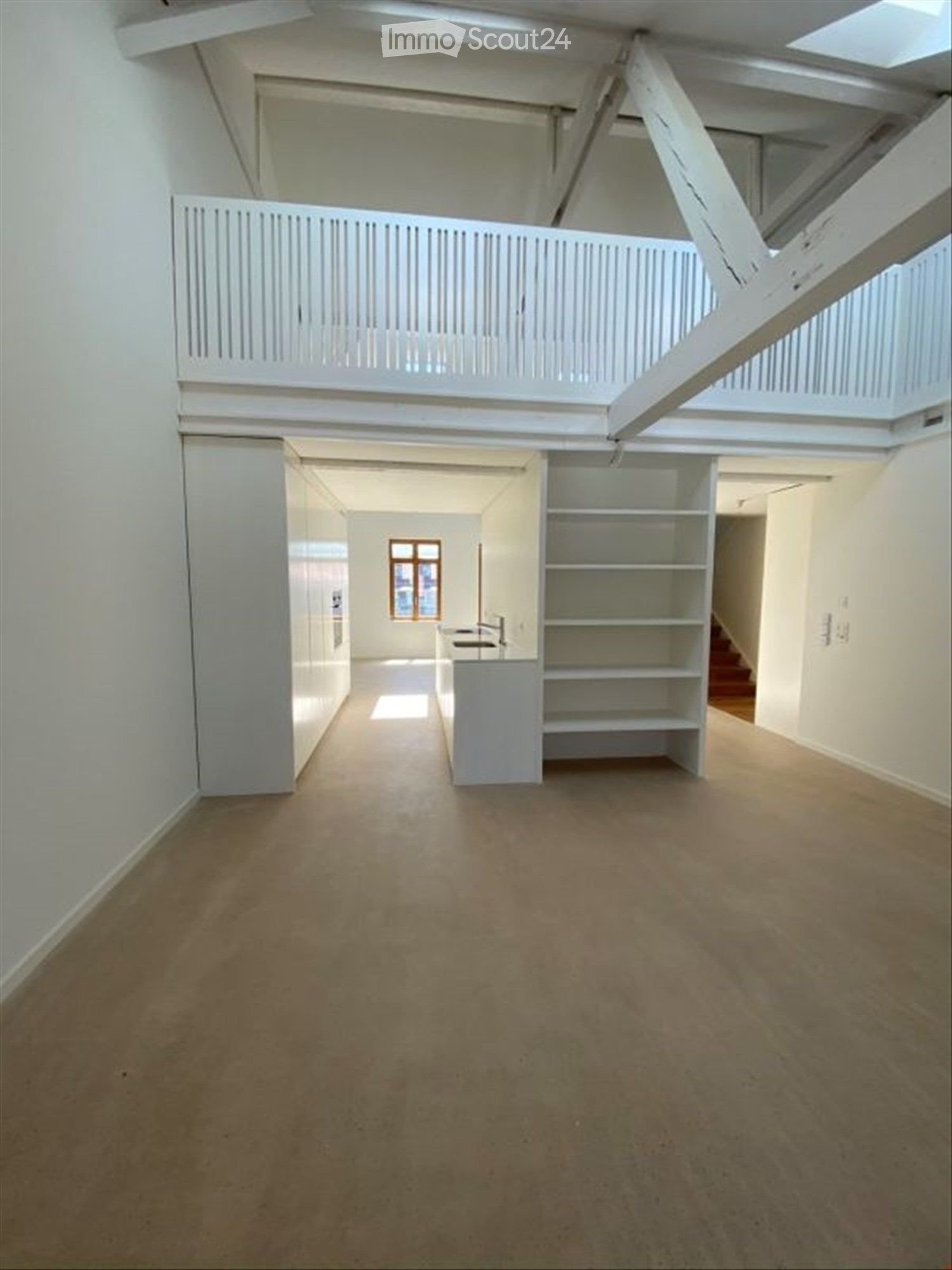 Empty room with white walls, concrete floor, and a small kitchen area with a sink and shelves. A staircase leads to an upper level with a white railing.