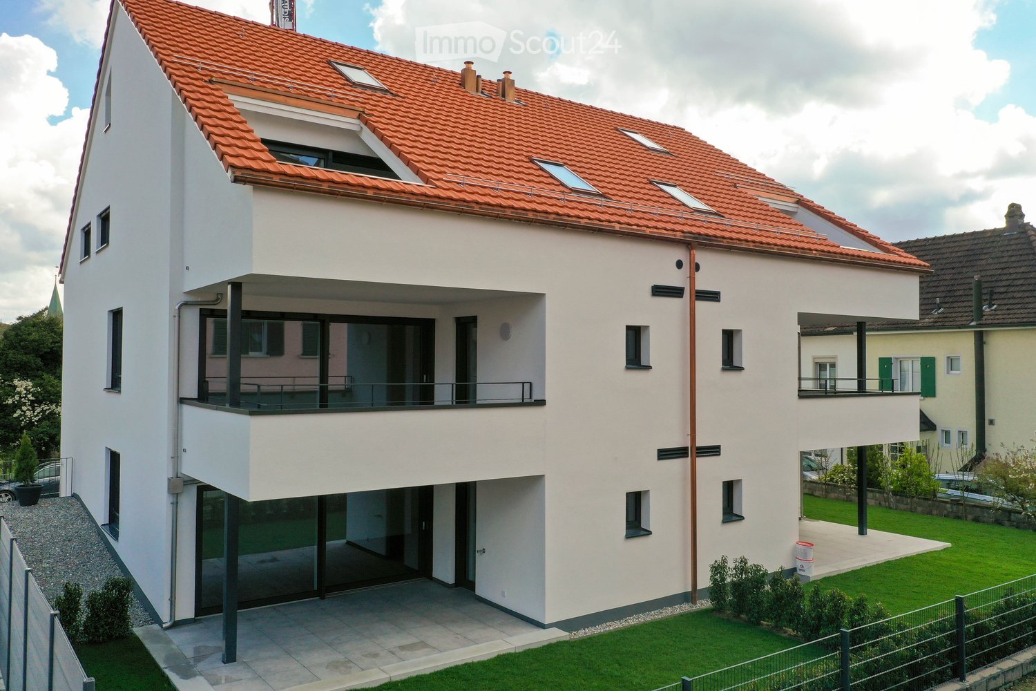 Modern two-story house with white exterior, red-tiled roof, multiple windows, and balconies. Features a raised ground floor, garage, and well-maintained garden with green lawn and small plants.