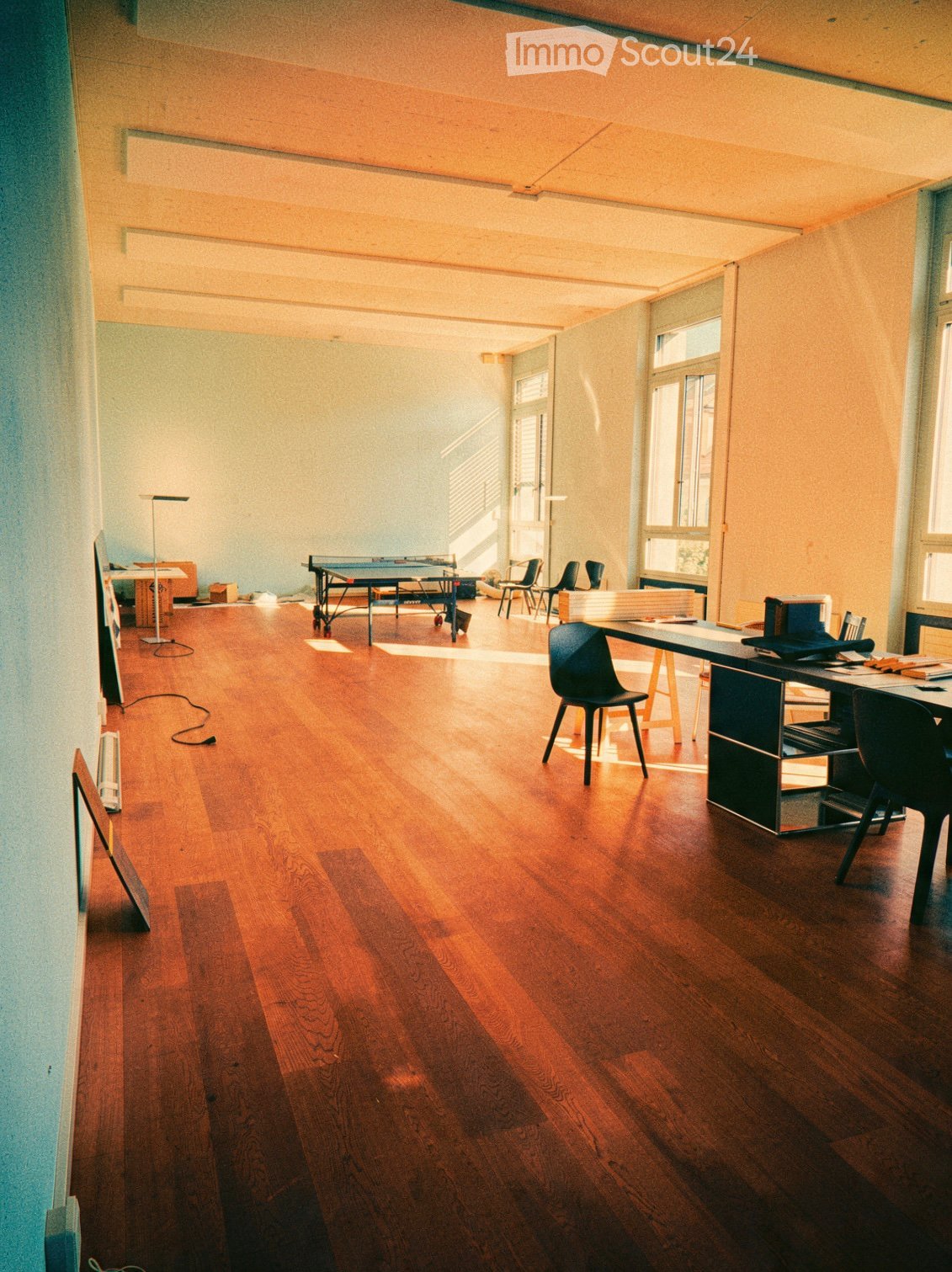Open, well-lit room with wooden flooring, a ping pong table, a desk with chairs, and a screen on the wall.