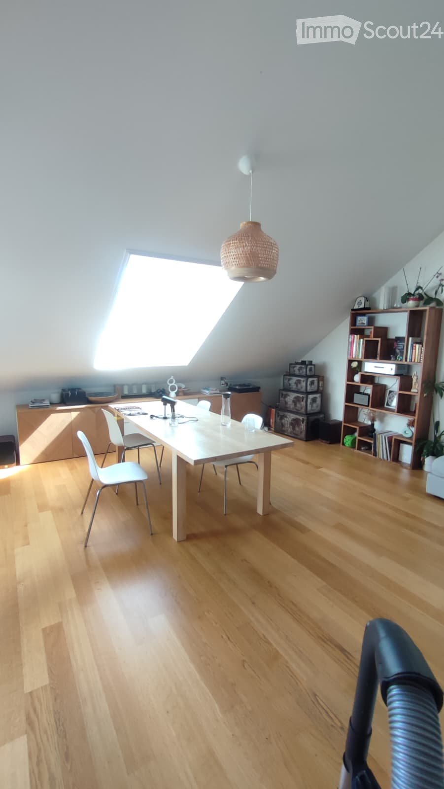 Bright attic room with wooden flooring, skylight window, dining table with chairs, bookshelf, and decorative items.