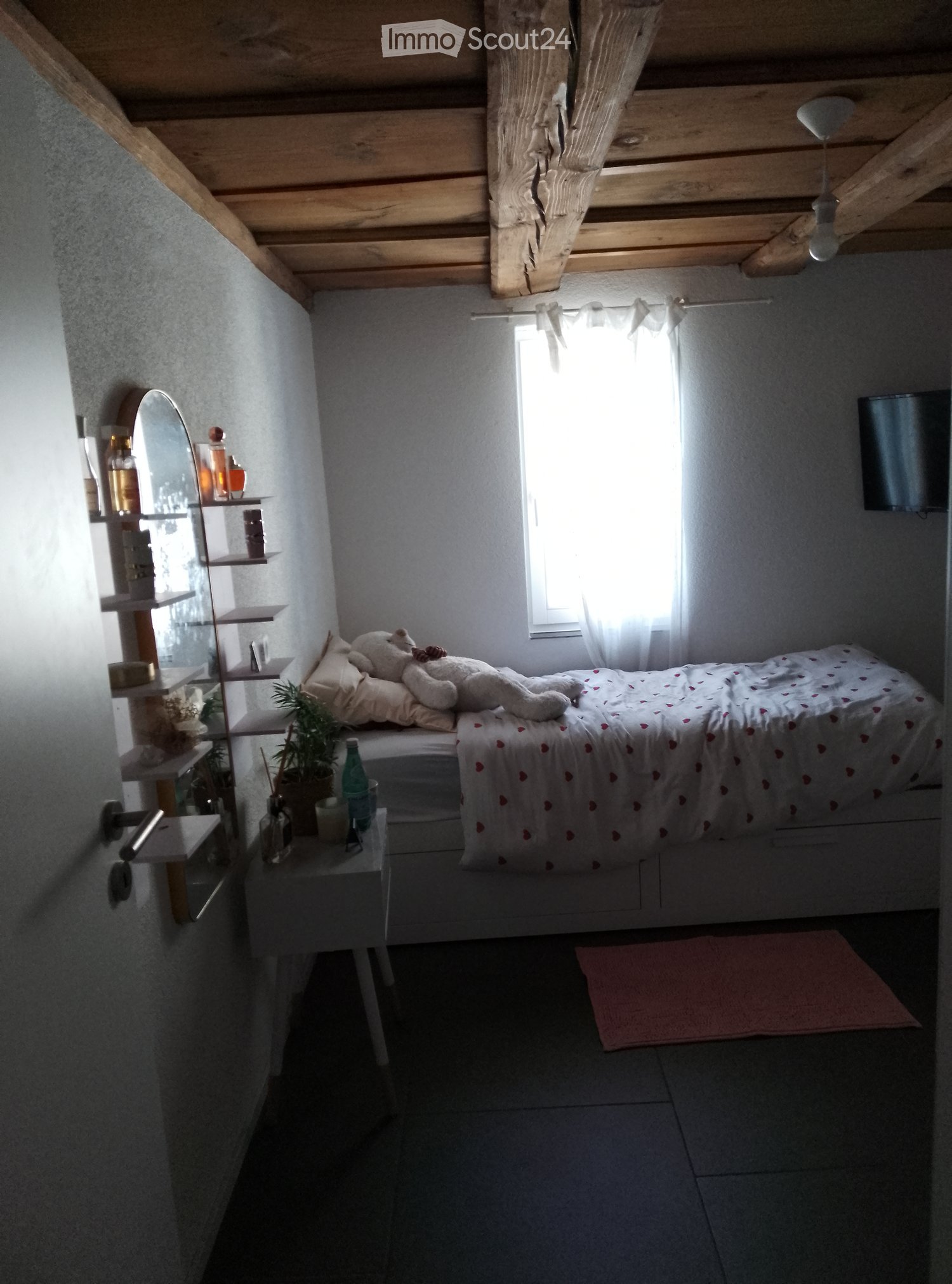 Cozy bedroom with a wooden slanted ceiling, a bed with a white comforter, a nightstand, a shelf with various items, and a small plant on the nightstand.