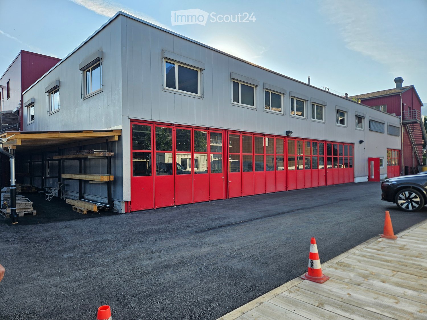 Modern two-story building with grey siding, red garage doors, and multiple windows. Includes a covered parking area and traffic cones on the asphalt ground.