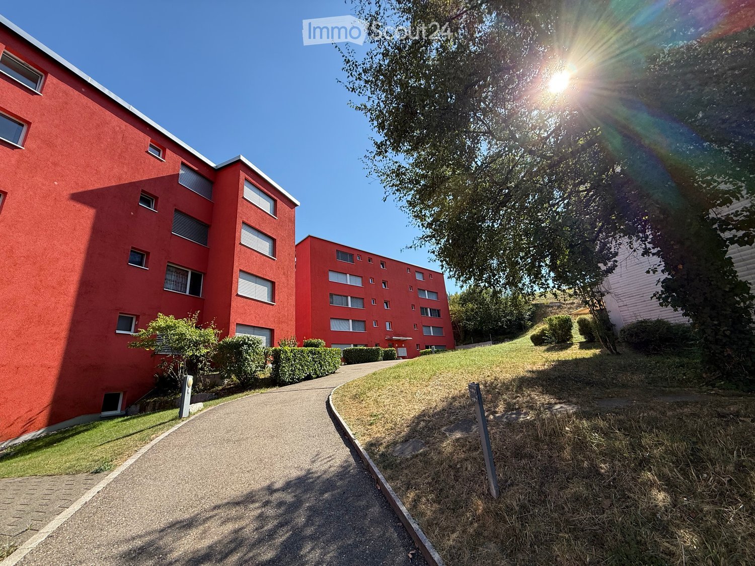Red apartment building with multiple windows and balconies, surrounded by greenery and a paved pathway.