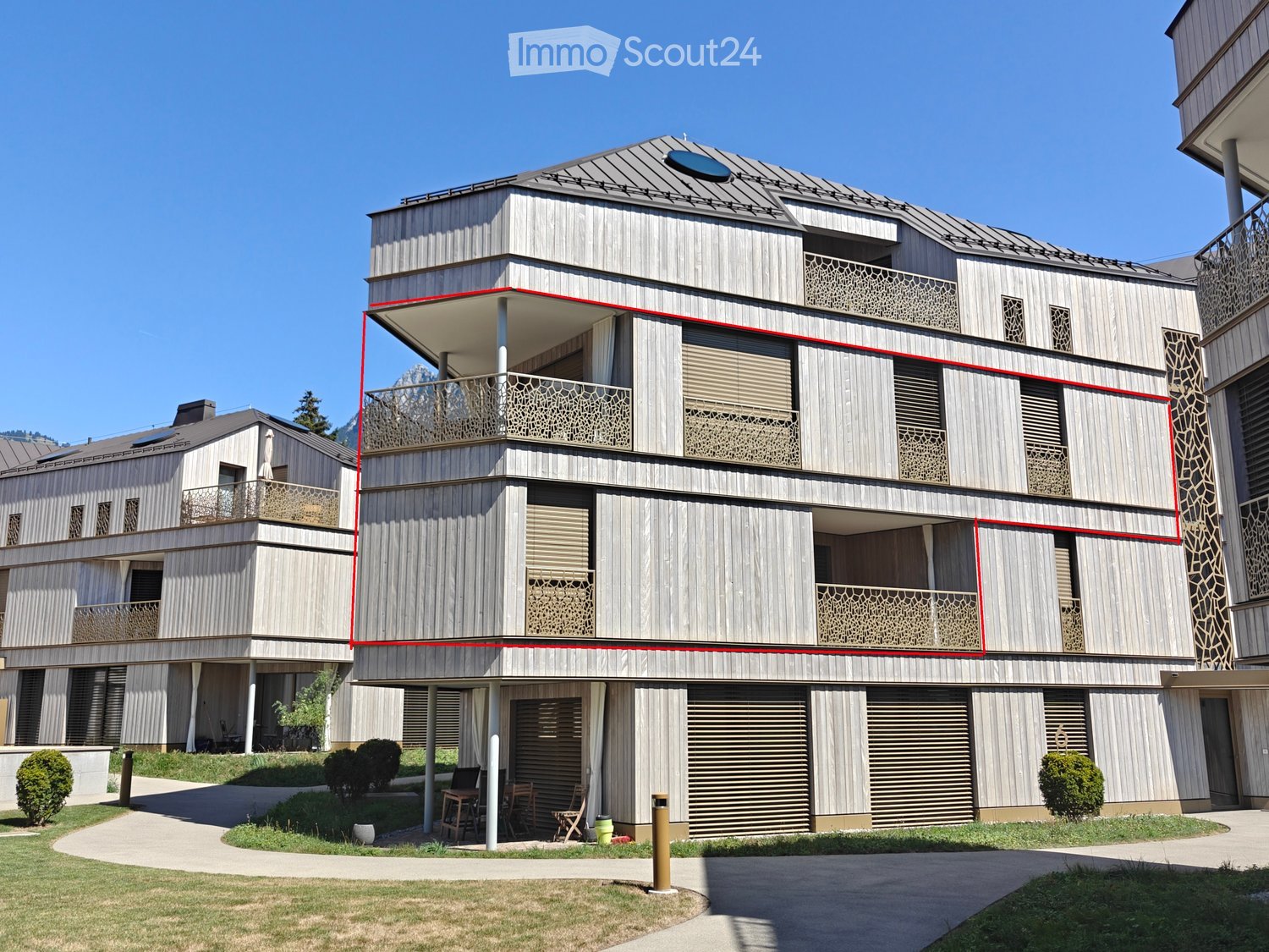Modern multi-story house with wooden exterior, balconies, and contemporary design. Features include large windows, decorative railings, and a flat roof with solar panels.