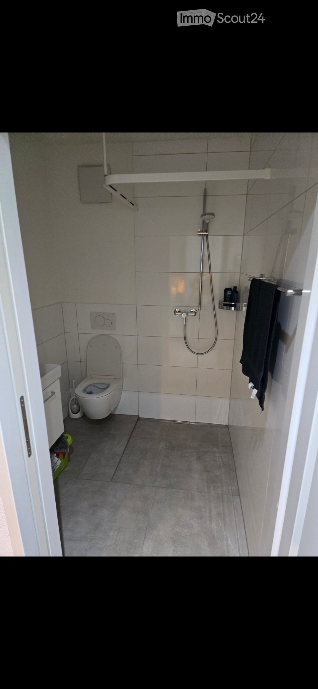 Modern bathroom with a toilet, shower, towel, and shower shelf. The walls are tiled in a light gray color, and the floor is covered with dark gray tiles.
