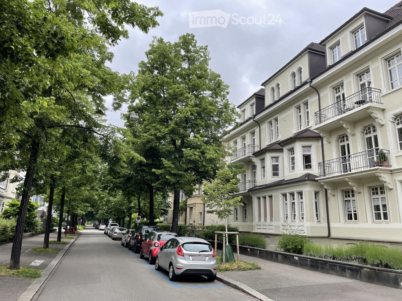 A multi-story residential building with balconies and large windows, situated on a tree-lined street with parked cars.