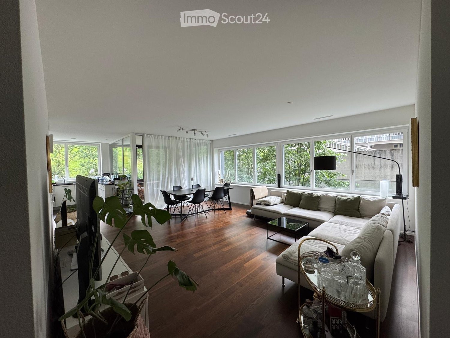 Modern living room with large windows, white sofa, coffee table, dining area, and a television. The room features wooden flooring and ample natural light.