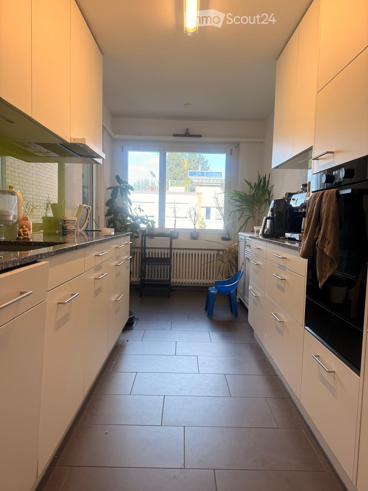 Modern kitchen with white cabinets, granite countertops, and a large window overlooking a balcony. Appliances include a sink, fridge, oven, and coffee maker. A small blue stool is placed near the window, and potted plants are on the windowsill.