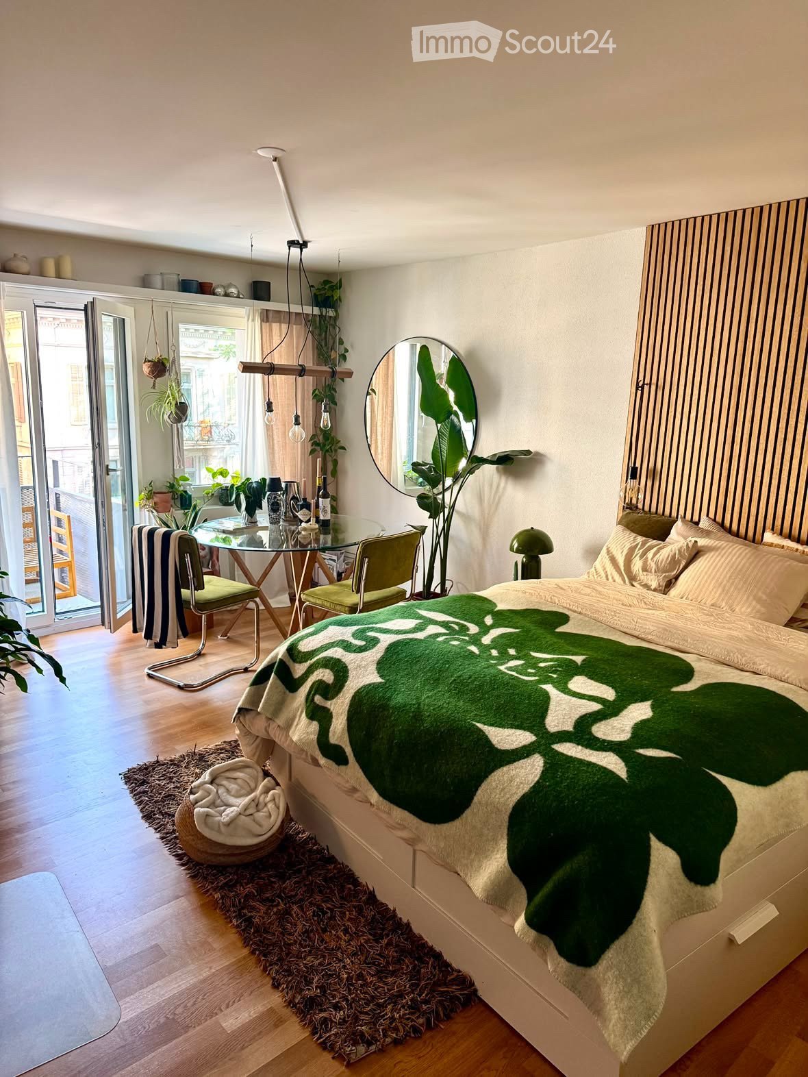 A modern bedroom with a large bed, green throw blanket, round mirror, hanging plants, and a small dining area with a glass table and chairs.