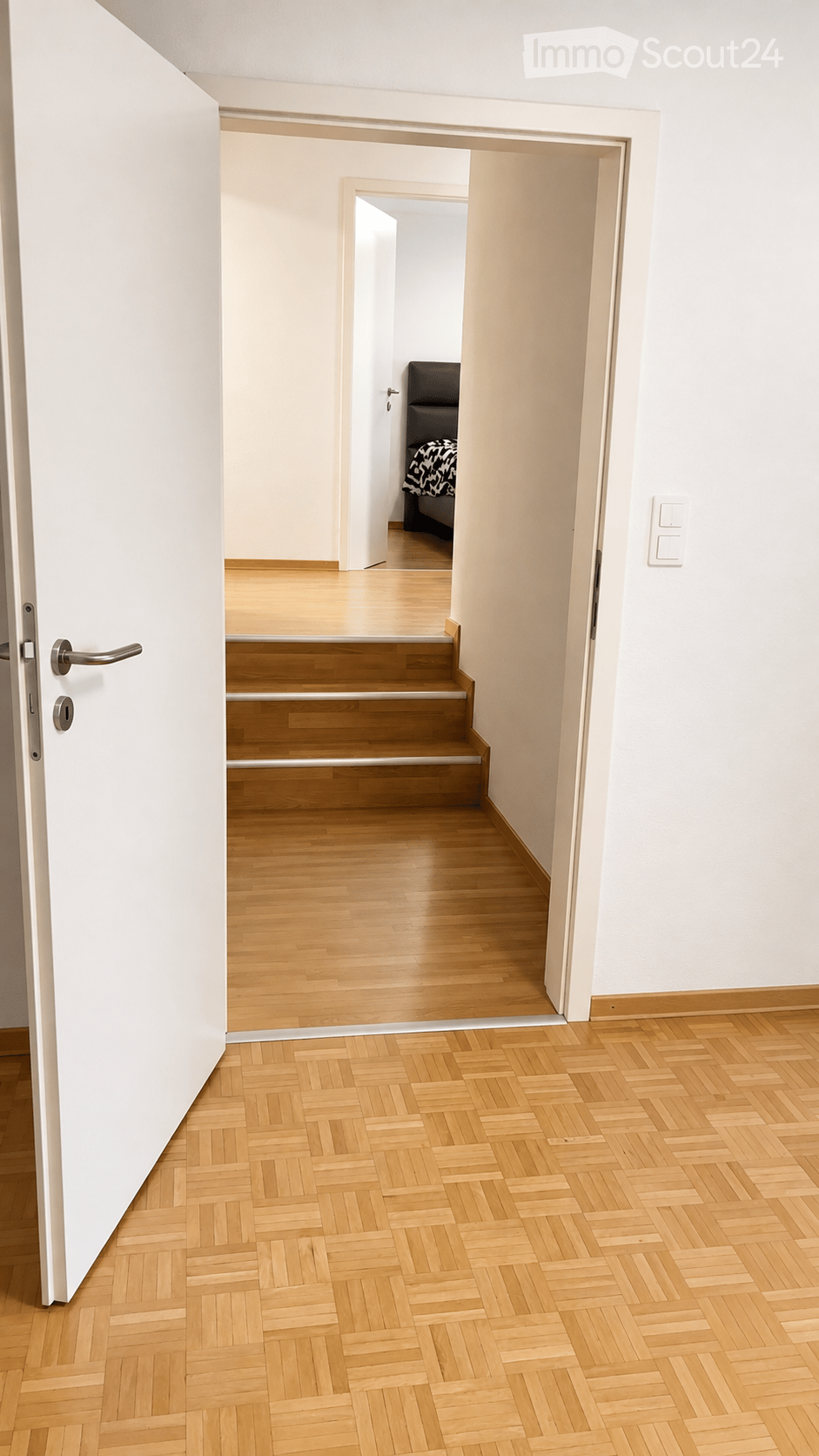 A clean, modern hallway with light wooden flooring and white walls. The hallway features a staircase leading upwards, with a door on the left and a glimpse of a bedroom visible at the end of the corridor.