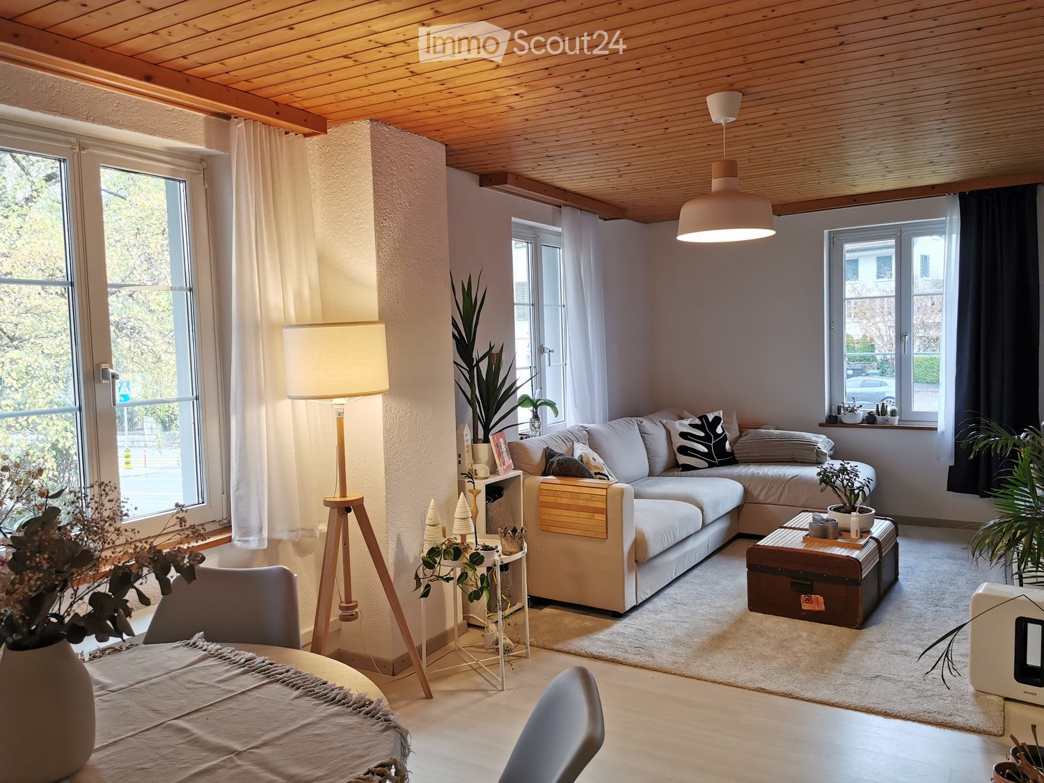 Modern living room with a white sectional sofa, coffee table, floor lamp, and large windows. Features a wooden ceiling and neutral color palette.