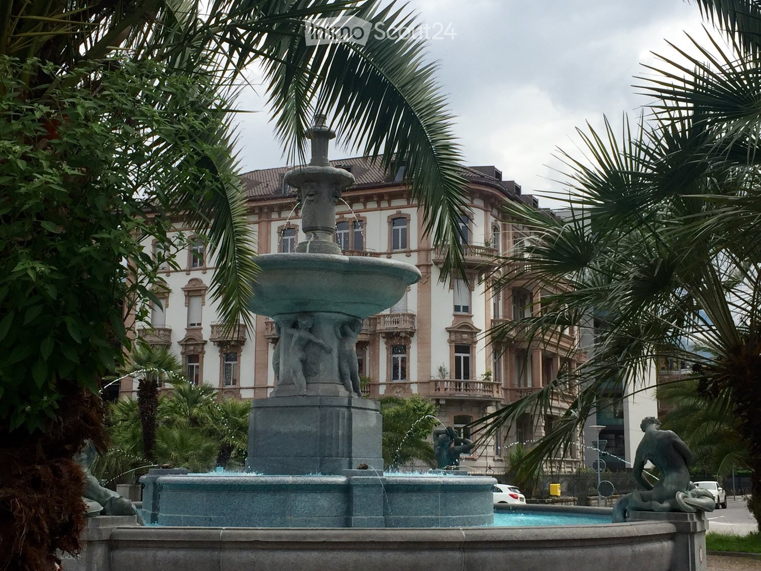 A large stone fountain with multiple tiers and sculptures, surrounded by palm trees and a grand historic building in the background.