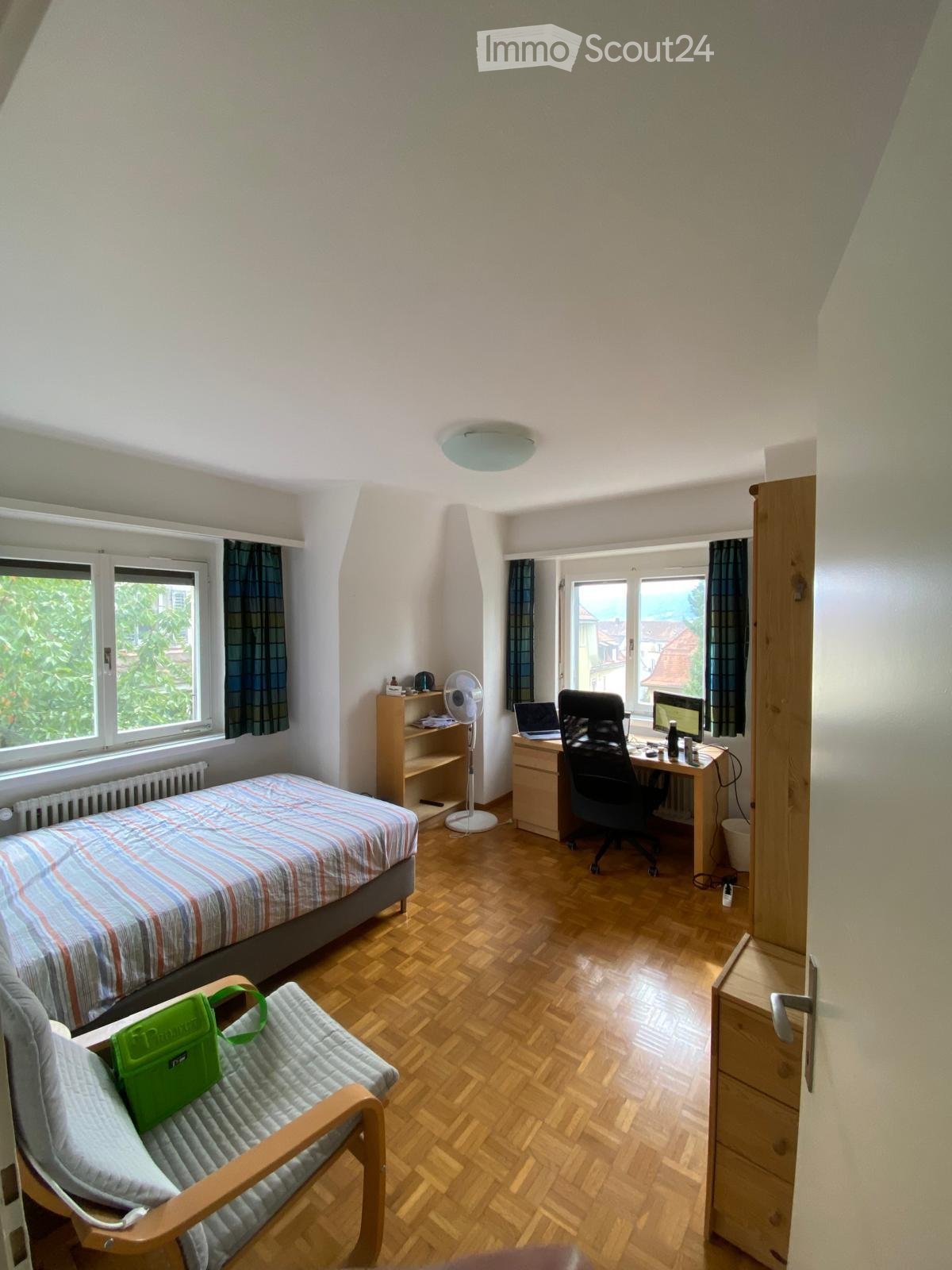 A furnished bedroom with a bed, desk, chair, laptop, fan, and a small chair with a green bag on it. The room has wooden flooring and large windows with curtains.