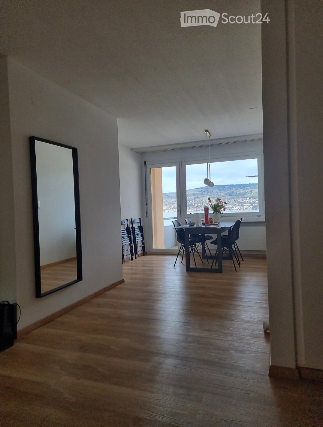 Bright, open dining area with wooden flooring, large windows offering a scenic view, a dining table with chairs, and a decorative flower vase on the table.