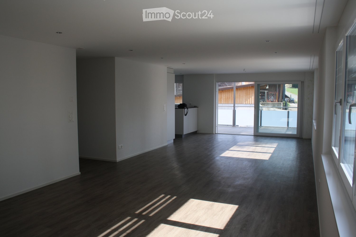 Empty room with wooden flooring, white walls, large windows, and a sliding glass door leading to an outdoor area.