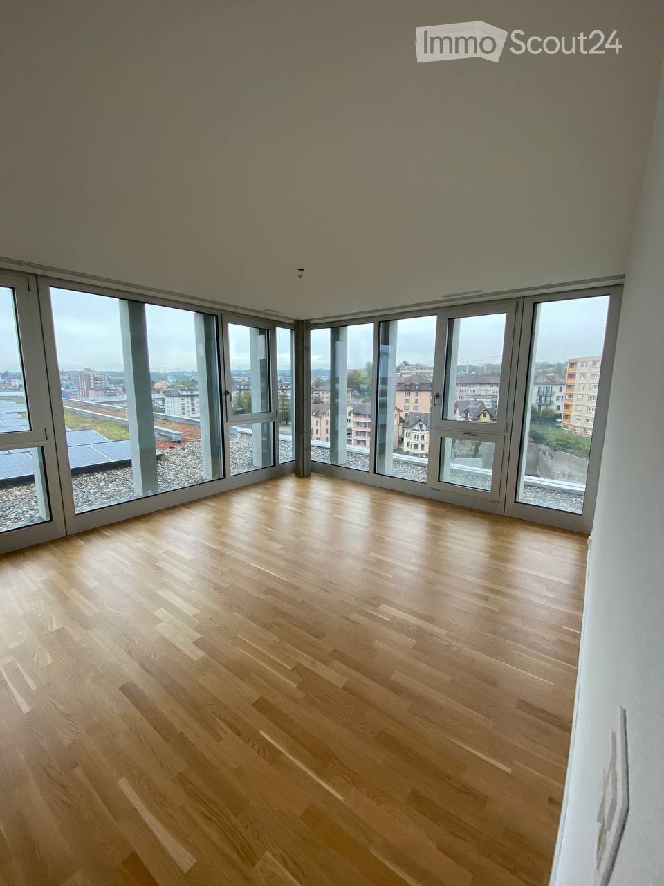 Completely empty room with wooden flooring and large windows offering a cityscape view.