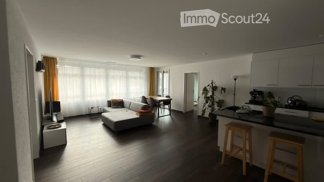 Open-plan living and dining area with a grey sectional sofa, coffee table, dining table with chairs, kitchen island with stools, and a potted plant. The space features large windows with sheer curtains and wooden flooring.