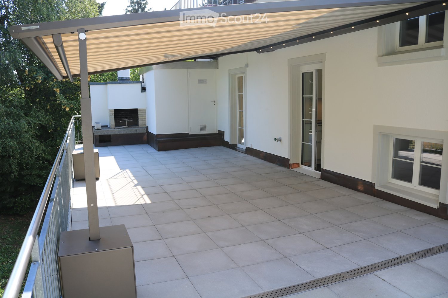 Balcony with tiled floor, metal railing, covered patio area with a retractable awning, and a fireplace in the background.