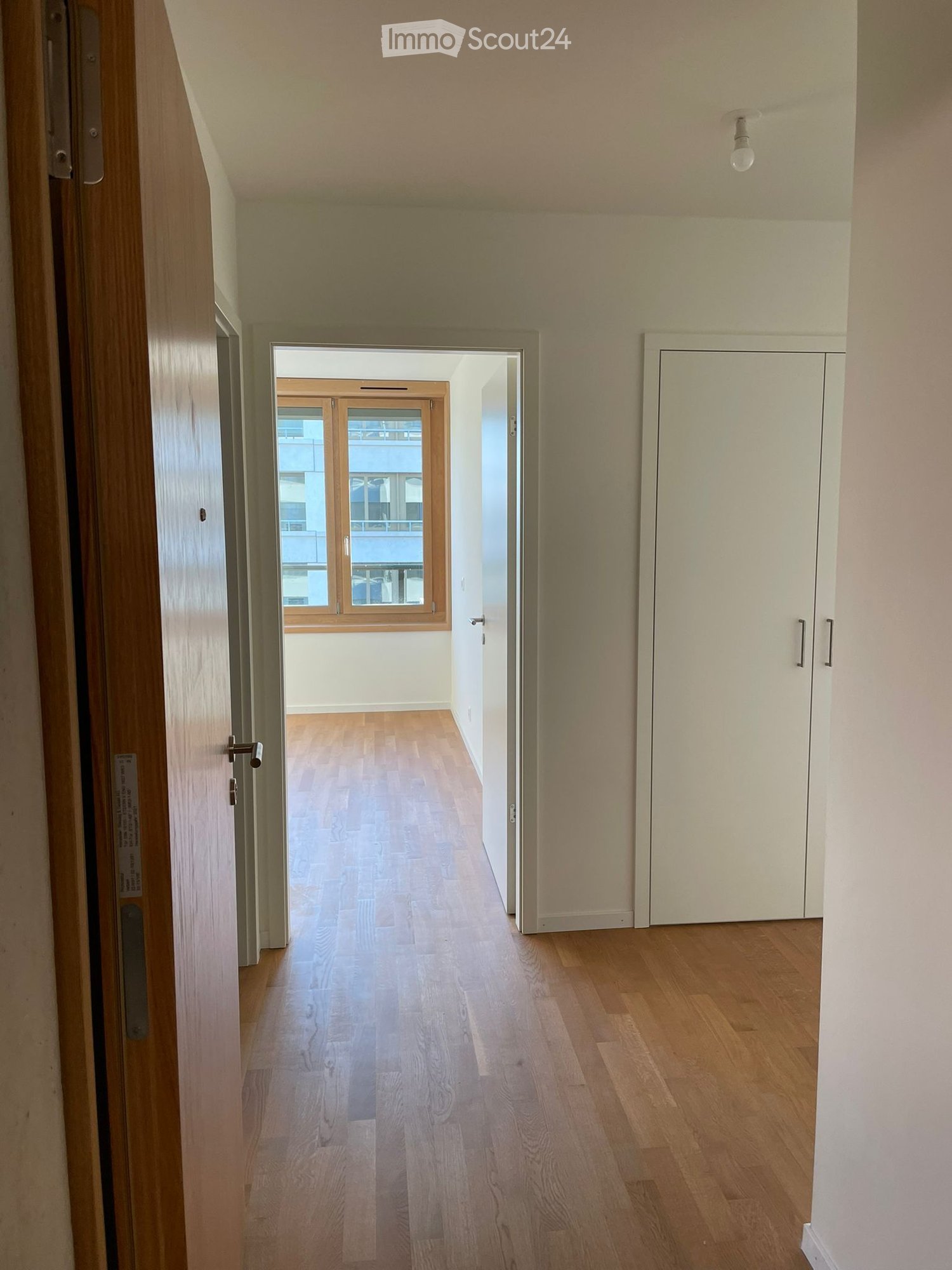 Empty hallway with wooden flooring, white walls, and a closed white cabinet on the right side. A door at the end leads to a room with large windows and a view of the outside.