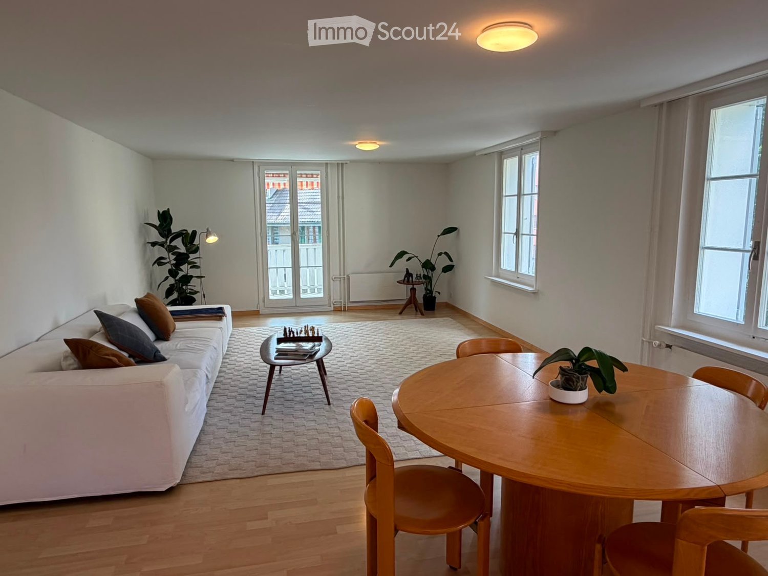 Bright living room with white walls, wooden flooring, large windows, and a balcony door. Features a white sofa with pillows, a coffee table with a chessboard, a dining table with chairs, and several potted plants.
