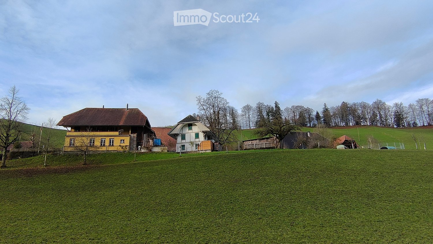 A two-story house with a yellow exterior and a brown roof, situated on a grassy field with trees and hills in the background.