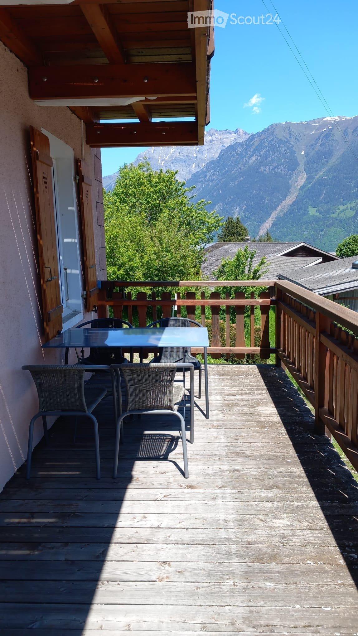 Balcony with wooden decking, glass table, and wicker chairs, overlooking a mountain view.
