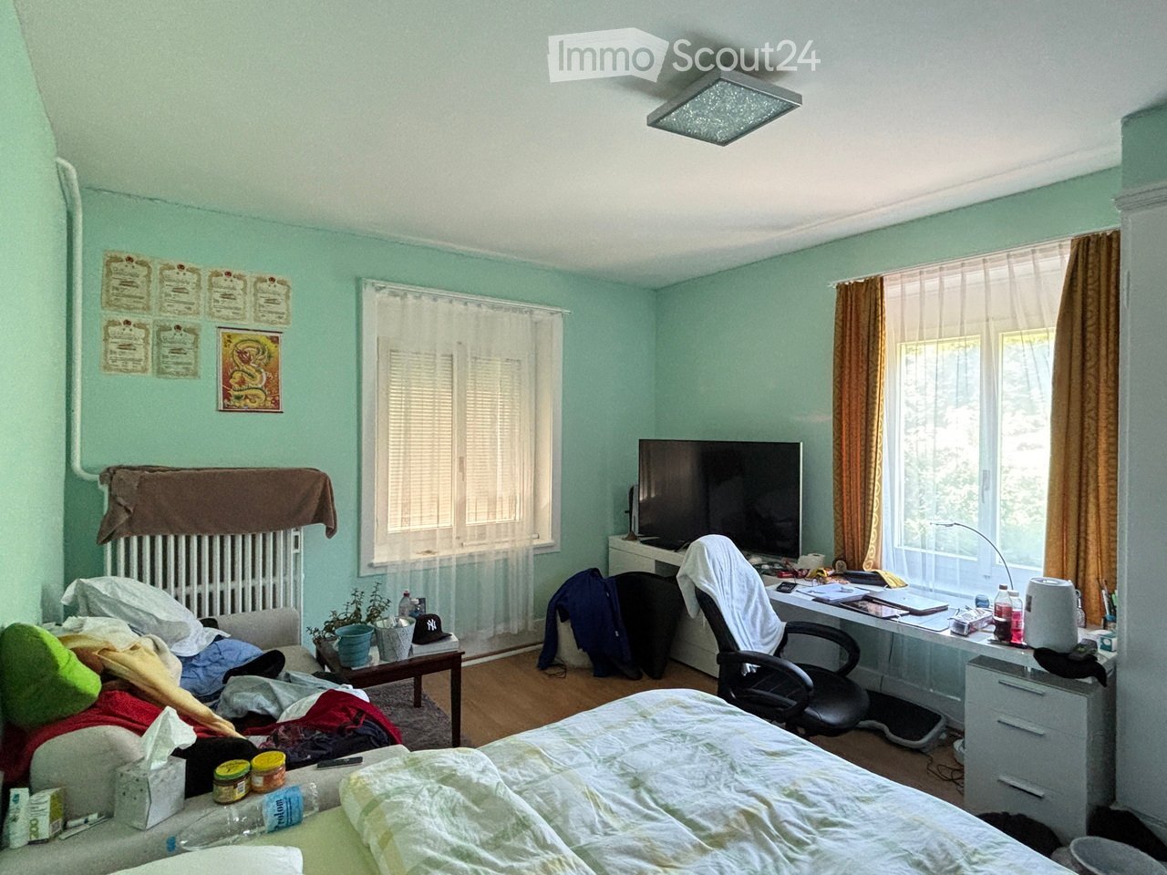 A bedroom with a bed, desk, chair, screen, small table, and various personal items. The room features light green walls, wooden flooring, and large windows with curtains.
