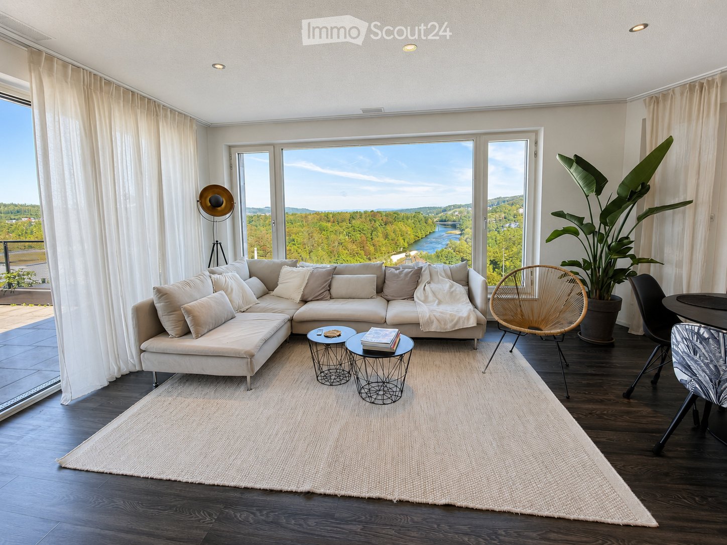 Modern living room with large windows offering a scenic view, featuring a sectional sofa, coffee tables, a wicker chair, and a large plant.