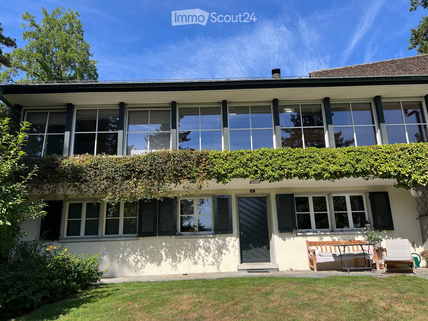 Two-story house with large windows, ivy-covered balcony, and outdoor seating area with a bench and chairs.