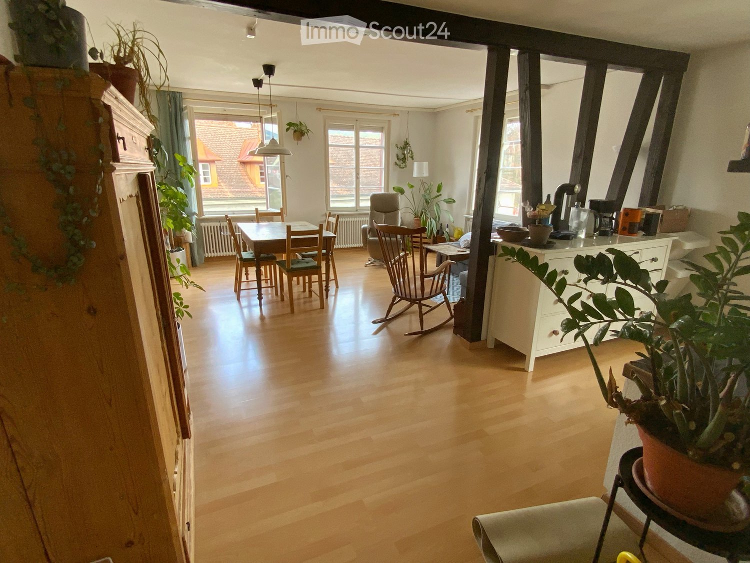Bright, open living and dining area with wooden flooring, large windows, a dining table with chairs, a rocking chair, and a white cabinet with potted plants.