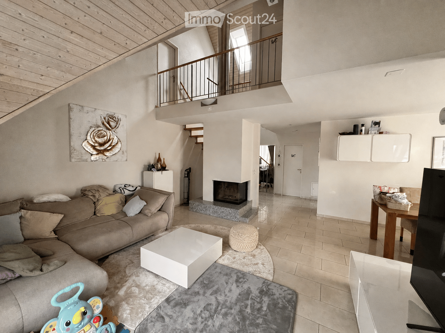 Modern living room with a beige sectional sofa, white coffee table, ottoman, fireplace, and a dining area with a table and chairs. The room features a large painting, a cabinet, and a staircase leading to an upper level.