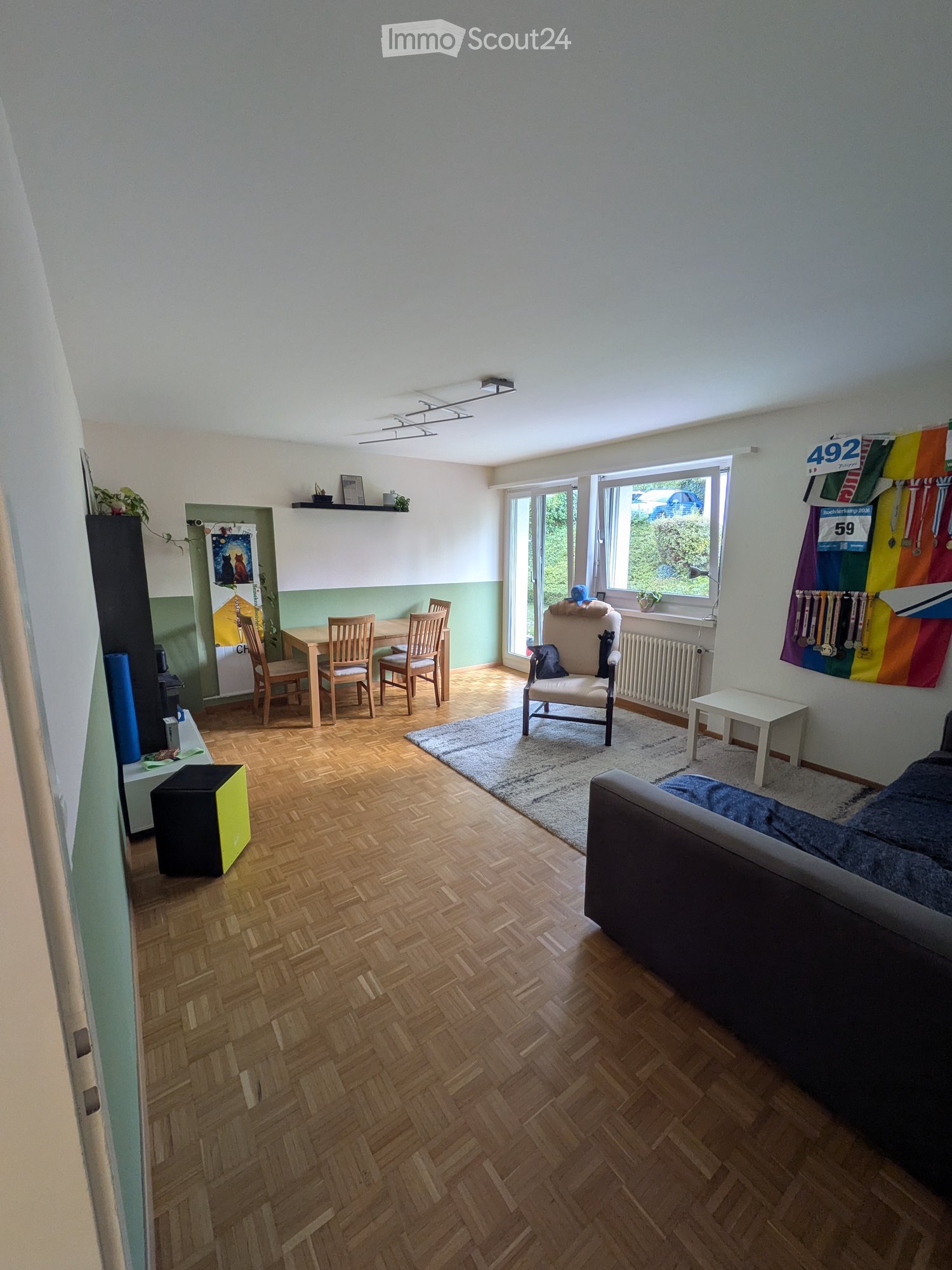 Living room with wooden flooring, large windows, sofa, armchair, dining table with chairs, small table, shelf with plants and picture frames, and a colorful banner on the wall.