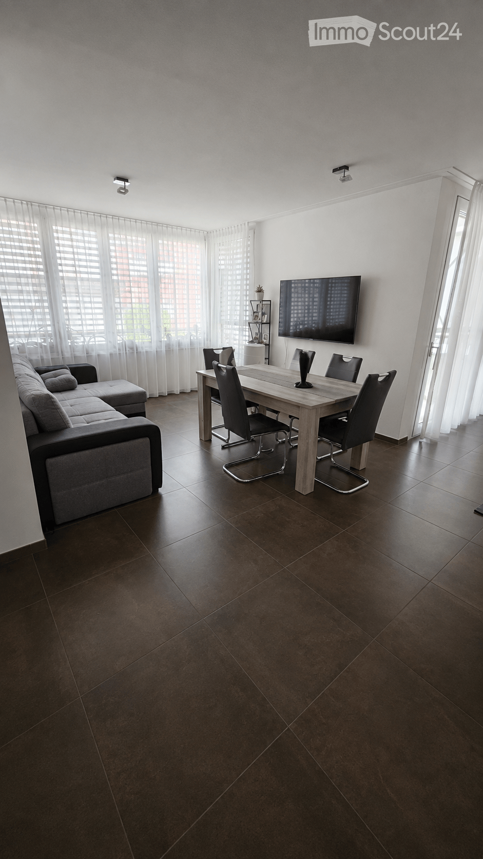 Modern living and dining area with dark brown tiled flooring, large windows with sheer curtains, a grey sectional sofa with an ottoman, a wooden dining table with four black chairs, a flat-screen TV mounted on the wall, and a small plant on a shelf.
