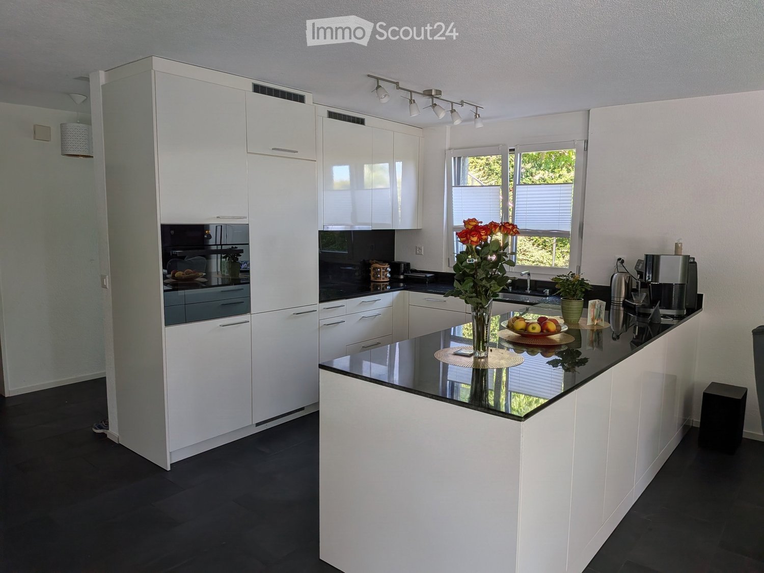 Modern kitchen with white cabinets, black countertops, and stainless steel appliances. Features a central island with a vase of flowers and a bowl of fruit, along with a coffee maker and a potted plant on the counter.